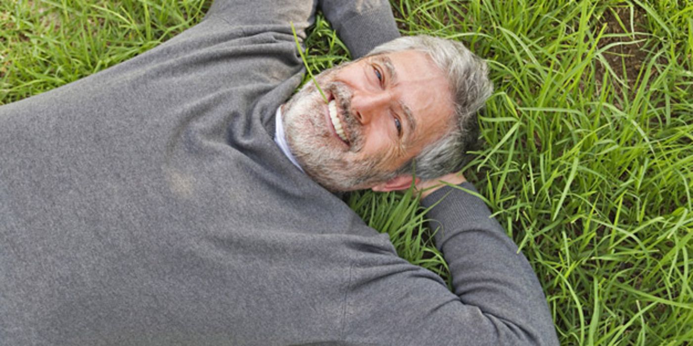 Foto von oben: Mann in den 50ern, lachend, im Gras liegend; graumelierter Bart und Haare, grauer V-Ausschnitt-Pullover, Arme hinter dem Kopf verschränkt