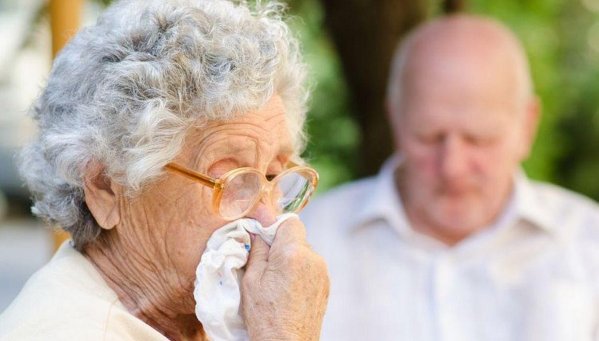 Senioren: Frau Vordergrund im Profil, grauhaarig, putzt sich die Nase, Hintergrund Mann und Natur, unscharf