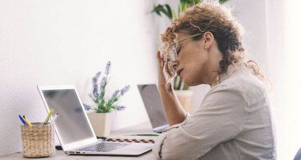 Frau fasst sich an den Kopf, während sie an einem Schreibtisch vor einem Laptop sitzt.