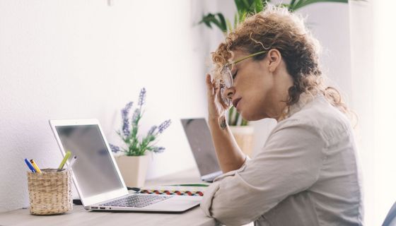 Frau fasst sich an den Kopf, während sie an einem Schreibtisch vor einem Laptop sitzt.