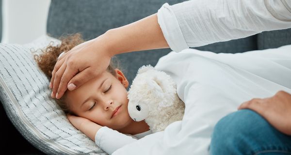 Krankes Kind liegt mit Kuscheltier auf dem Sofa und Mutter hält die Hand an die Stirn.