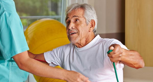 Älterer grauhaariger Mann in einem Gymnastikraum bei der Physiotherapie, den Anweisungen der Therapeutin lauschend