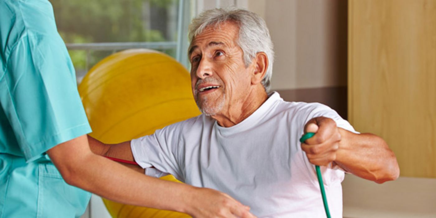 Älterer grauhaariger Mann in einem Gymnastikraum bei der Physiotherapie, den Anweisungen der Therapeutin lauschend
