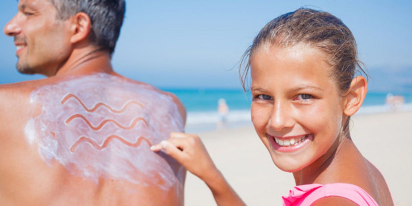 Strandszene, Mann, Halbprofil, Sonnencreme auf Rücken, die von einem ca. 14 Jährigen Mädchen, das in die Kamera lacht in Zickzackmuster gemalt wird