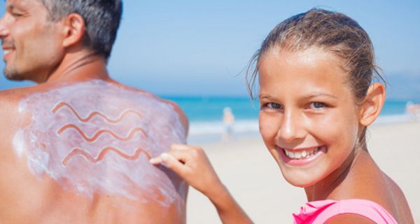 Strandszene, Mann, Halbprofil, Sonnencreme auf Rücken, die von einem ca. 14 Jährigen Mädchen, das in die Kamera lacht in Zickzackmuster gemalt wird