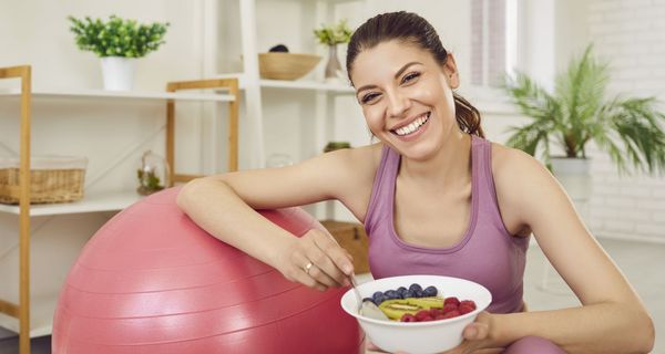 Junge Frau mit Früchteteller in Sportkleidung, einen Arm auf einem Gymnastikball.