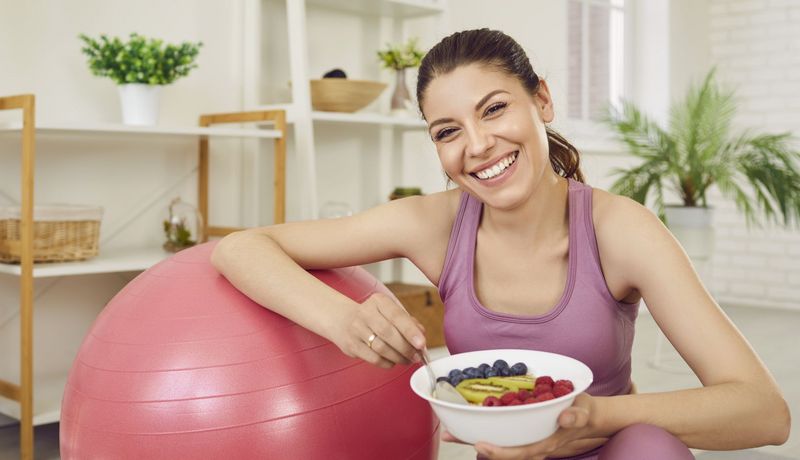 Junge Frau mit Früchteteller in Sportkleidung, einen Arm auf einem Gymnastikball.