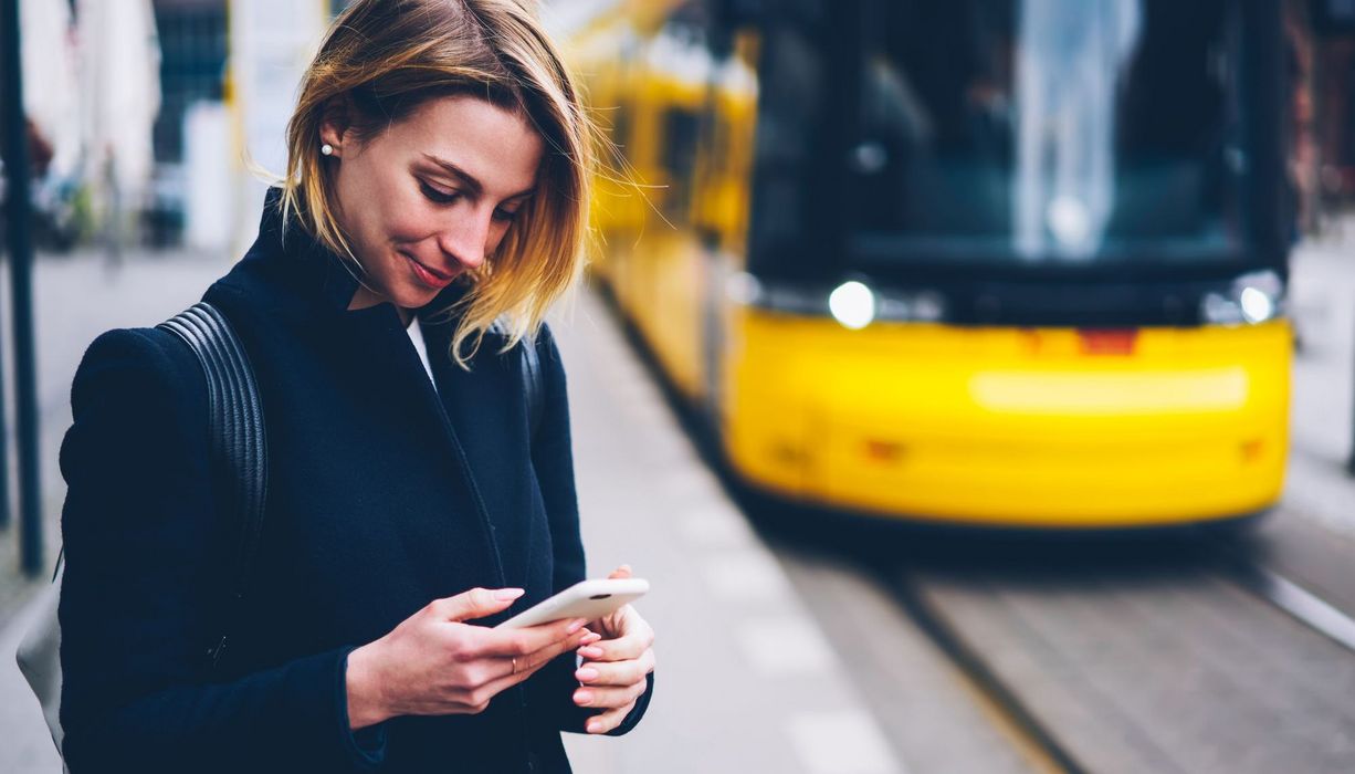 Junge Frau, steht an der Straßenbahnhaltestelle mit ihrem Smartphone.