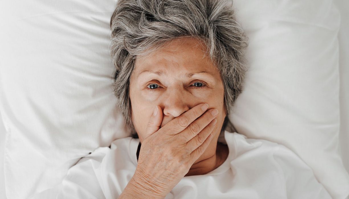 Ältere Frau, liegt im Bett und hält sich eine Hand vor den Mund. 