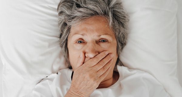 Ältere Frau, liegt im Bett und hält sich eine Hand vor den Mund. 