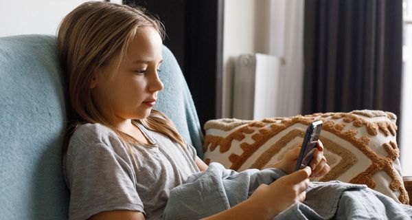Ein junges Mädchen sitzt auf einem Sofa und hält lächelnd ein Smartphone in der Hand.