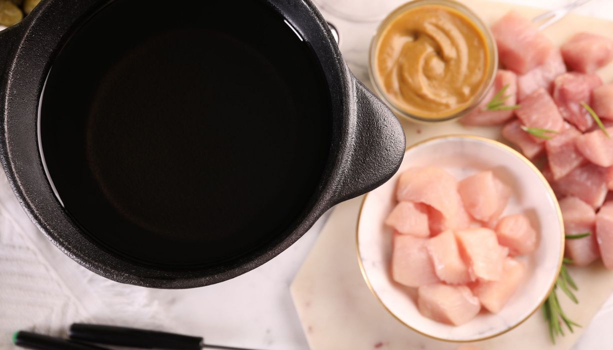 Fonduetopf mit Öl von oben, daneben rohes Hühnerfleisch, Schweinefleisch, Senf und Fondue-Gabeln.