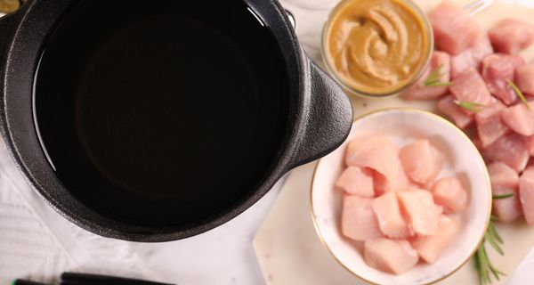Fonduetopf mit Öl von oben, daneben rohes Hühnerfleisch, Schweinefleisch, Senf und Fondue-Gabeln.