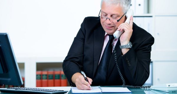 Älterer Mann im Anzug sitzt am Schreibtisch, telefoniert und notiert sich etwas