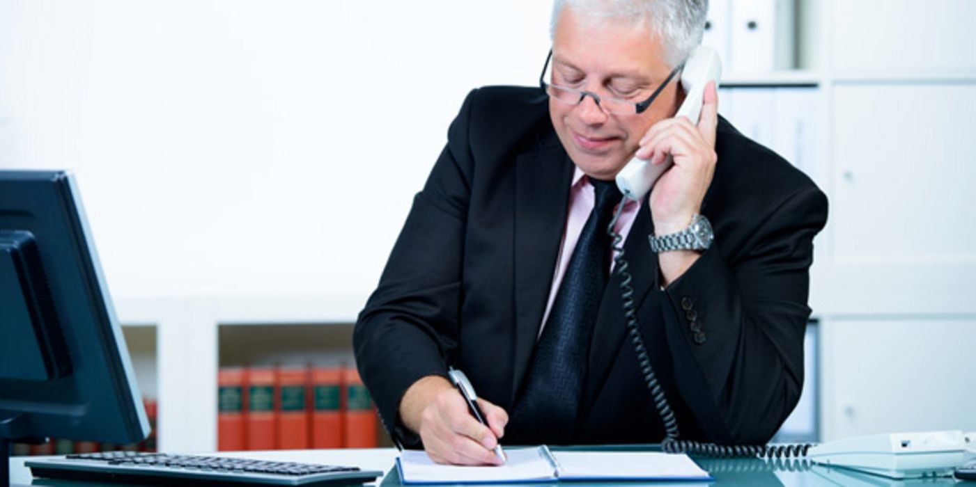 Älterer Mann im Anzug sitzt am Schreibtisch, telefoniert und notiert sich etwas