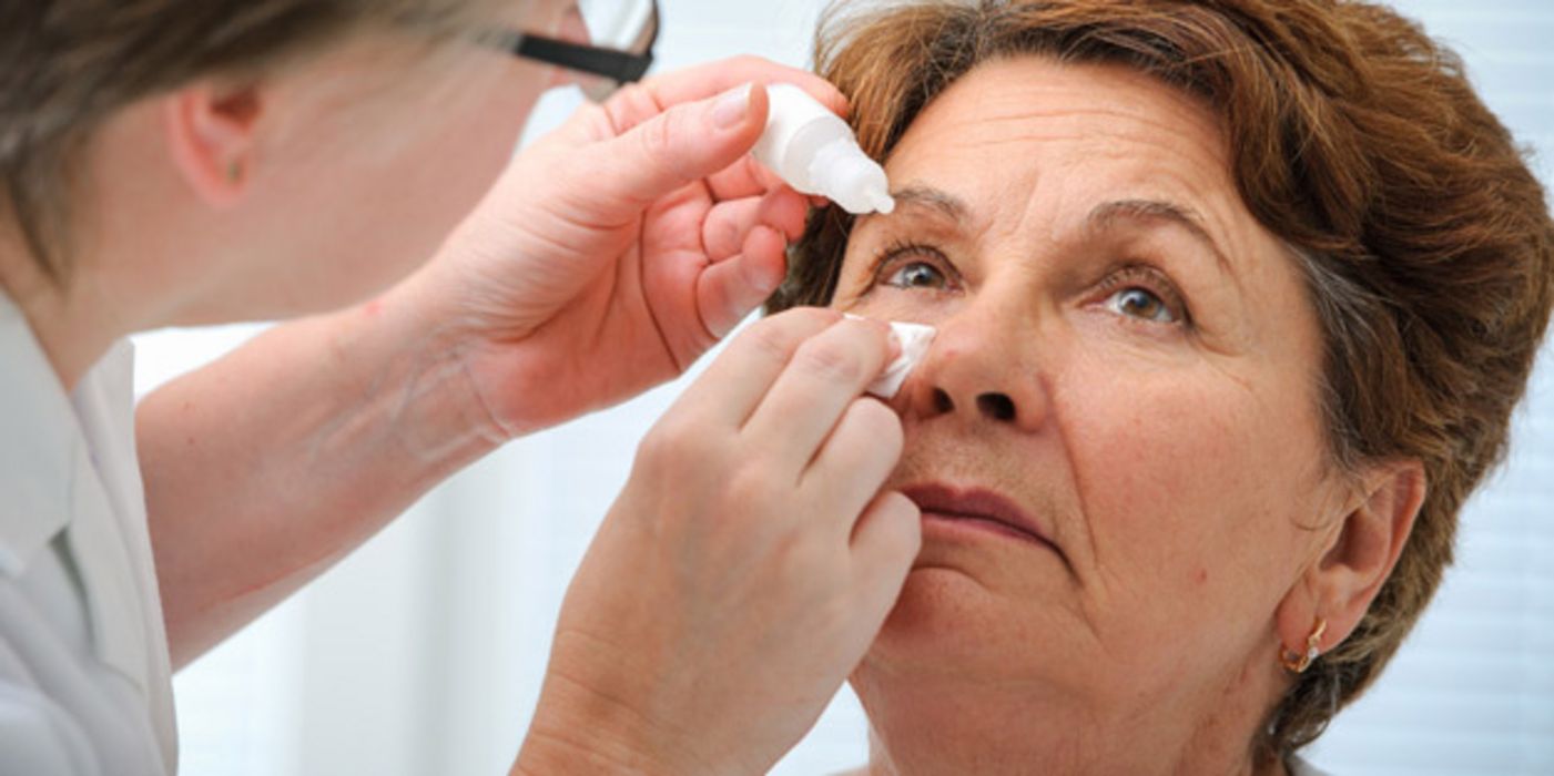 Nahaufnahme Gesicht: Ältere Frau bekommt von Ärztin (schemenhaft am Bildrand zu erkennen) Augentropfen verabreicht