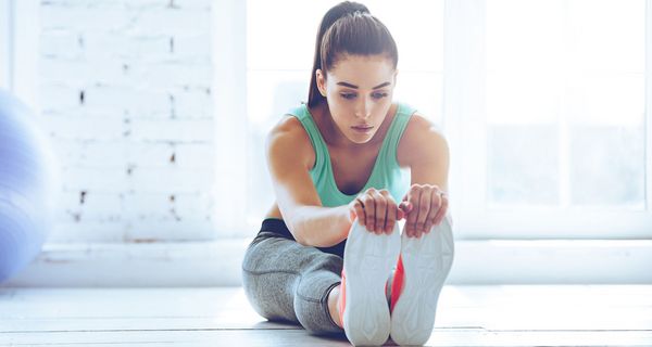 Junge Frau mit Sportkleidung, dehnt ihre Beine, indem sie sich mit dem Oberkörper nach vorn zu den Füßen beugt.