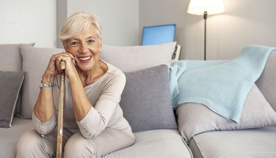 Ältere Seniorin, sitzt auf der Couch, stützt sich auf ihren Gehstock und lächelt in die Kamera.