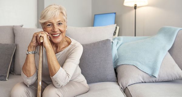 Ältere Seniorin, sitzt auf der Couch, stützt sich auf ihren Gehstock und lächelt in die Kamera.