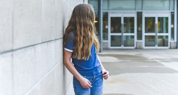 Junges Mädchen, steht vor der Schule und dreht der Kamera den Rücken zu.