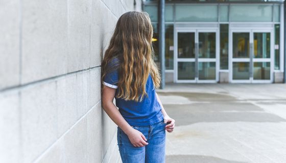 Junges Mädchen, steht vor der Schule und dreht der Kamera den Rücken zu.