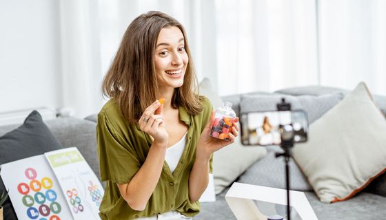 Frau nimmt sich mit Smartphone auf, während sie eine orangene Lutschpastille in die Kamera hält und lächelt. Hinter ihr lehnt eine Tafel zu B-Vitaminen.
