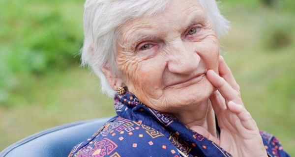 Porträtfoto: alte, grauhaarige Dame in blaugrundigem Morgenmantel, sitzend, schaut in die Kamera, Hintergrund Garten