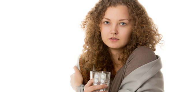 Junge Frau, dunkele, längere Locken, braune Decke um die Schultern, Wasserglas und Blisterpackung in der Hand