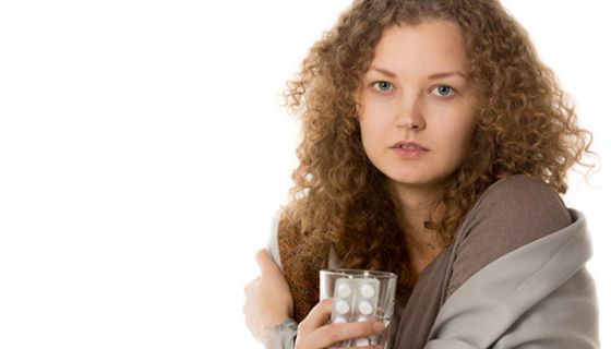 Junge Frau, dunkele, längere Locken, braune Decke um die Schultern, Wasserglas und Blisterpackung in der Hand