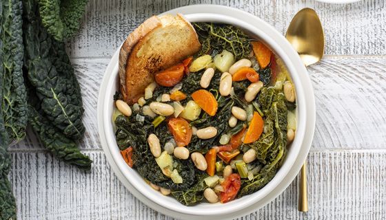 Draufsicht auf eine Schüssel mit Ribollita, toskanischer Gemüsesuppe mit Palmkohl und Brot. Daneben ein Löffel und einige Blätter des Palmkohls.