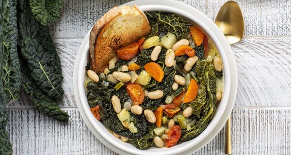 Draufsicht auf eine Schüssel mit Ribollita, toskanischer Gemüsesuppe mit Palmkohl und Brot. Daneben ein Löffel und einige Blätter des Palmkohls.