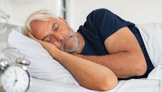 Mann, mindestens 50 Jahre alt, schläft in seinem Bett; im Vordergrund: Ein Wecker