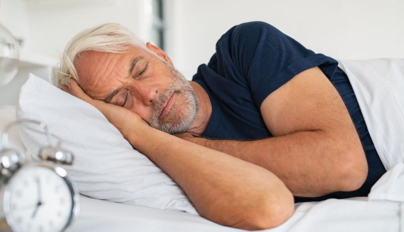 Mann, mindestens 50 Jahre alt, schläft in seinem Bett; im Vordergrund: Ein Wecker
