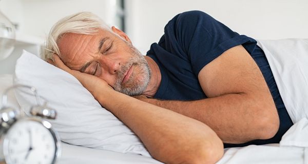 Mann, mindestens 50 Jahre alt, schläft in seinem Bett; im Vordergrund: Ein Wecker