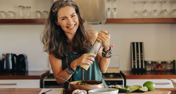 eine Frau um die 50 bereitet sich einen Salat in der Küche zu und lacht in die Kamera.