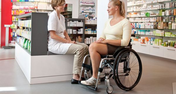 Apothekerin spricht mit junger Frau im Rollstuhl.