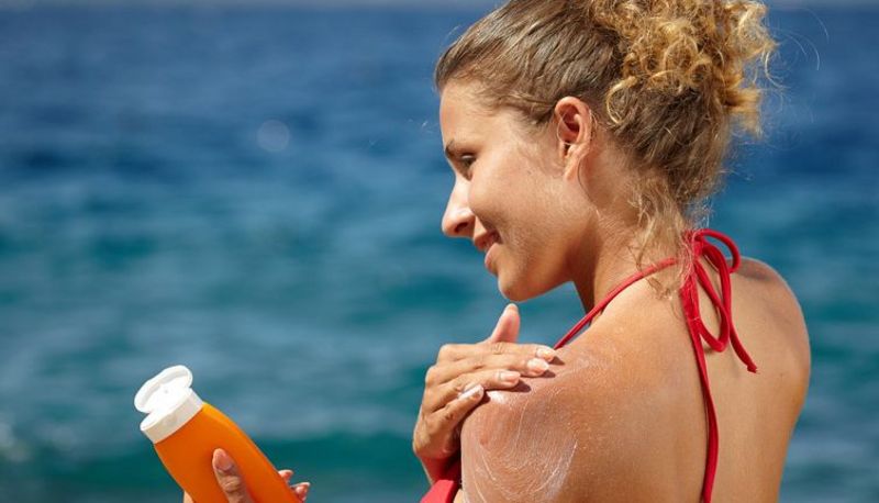 Jüngere Frau im roten Bikini, Rücken- und Schulteransicht, blonde, hochgebundene Haare, cremt sich mit Sonnenschutz die linke Schulter ein; Hintergrund: blaues Meer