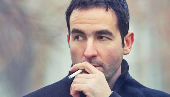 Porträtfoto: Mann, ca. Ende 30, dunkle, kurze Haare, dunkelgrauer Mantel (Ansatz zu sehen), Hintergrund unscharf, raucht und schaut nach links aus dem Bild heraus