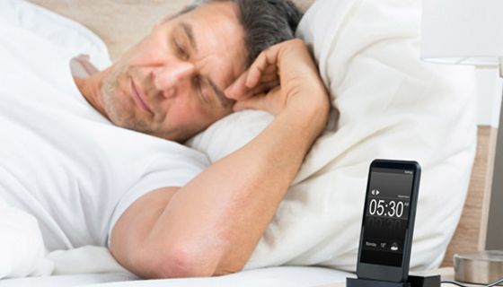 Mann um die 40 schläft auf der Seite im weißbezogenen Bett, weißes T-Shirt, Vordergrund Nachttisch mit Smartphone in Ladestation