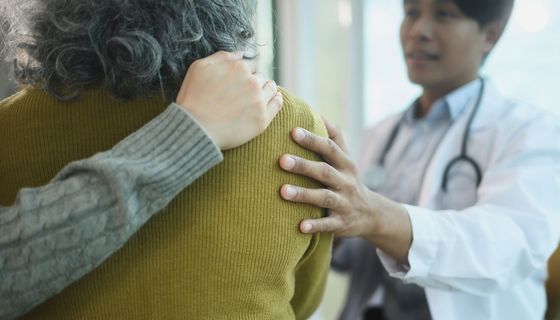 Blick in den Behandlungsraum auf die Rücken einer älteren Patientin von hinten: Ihre Begleitung hat einen Arm um sie gelegt, der Arzt hält ihr tröstend die Schulter. 