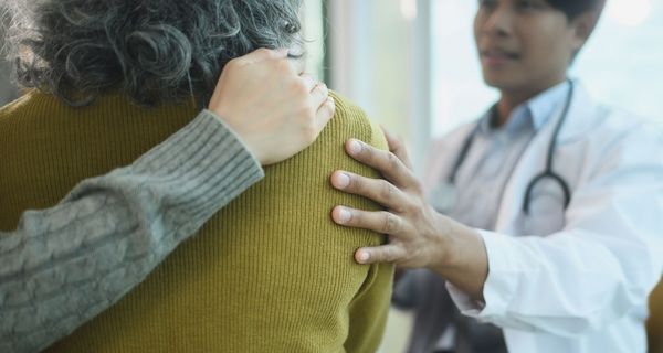 Blick in den Behandlungsraum auf die Rücken einer älteren Patientin von hinten: Ihre Begleitung hat einen Arm um sie gelegt, der Arzt hält ihr tröstend die Schulter. 