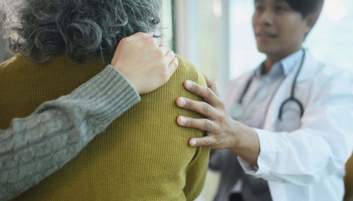 Blick in den Behandlungsraum auf die Rücken einer älteren Patientin von hinten: Ihre Begleitung hat einen Arm um sie gelegt, der Arzt hält ihr tröstend die Schulter. 