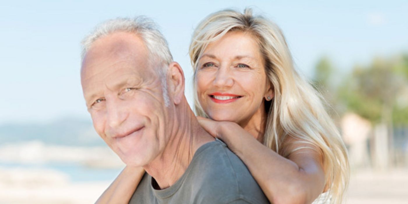Portrait (bis Schultern) attraktives Paar, sommerlich, er ca. Mitte 60, sie Mitte 50 mit grauem, langem Haar, vor schemenhaftem Strandhintergrund