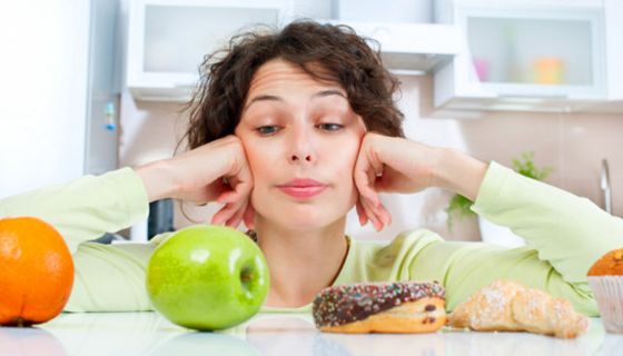 Frau, vor der Orange, Apfel, Donut, Croissant und Muffin aufgereiht sind und die unglücklich auf den Apfel blickt