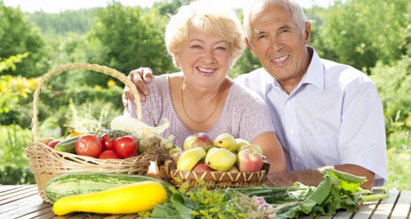 Fröhliches Seniorenpaar im Sommer im Freien an einem Tisch mit Obst und Gemüse, z.T. in Körben, z.T. auf dem Tisch liegend