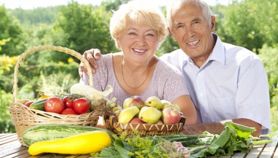 Fröhliches Seniorenpaar im Sommer im Freien an einem Tisch mit Obst und Gemüse, z.T. in Körben, z.T. auf dem Tisch liegend