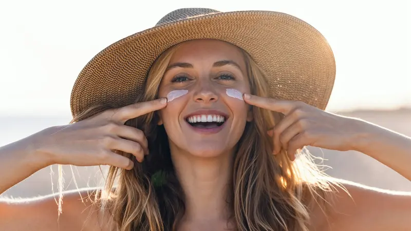 Frau mit Sonnenhut cremt sich das Gesicht ein.