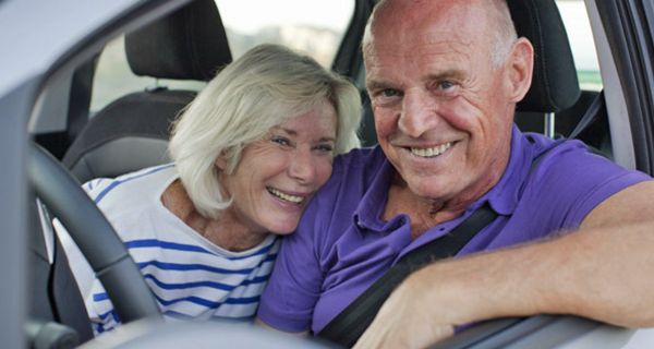 Paar um die 60, Mann am Steuer, lachen fröhlich aus dem offenen Fahrerfenster heraus in die Kamera, sie an die Schulter des Mannes gelehnt