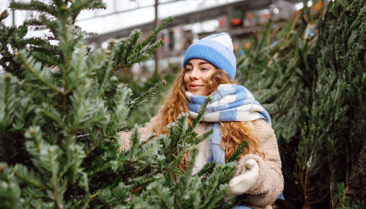 Eine Frau hockt zwischen vielen Weihnachtsbäumen. Sie sieht glücklich aus.