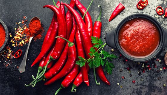 Rote Chilischoten, Chilipulver und eine Chilsoße auf einer schwerzen Tischplatte.